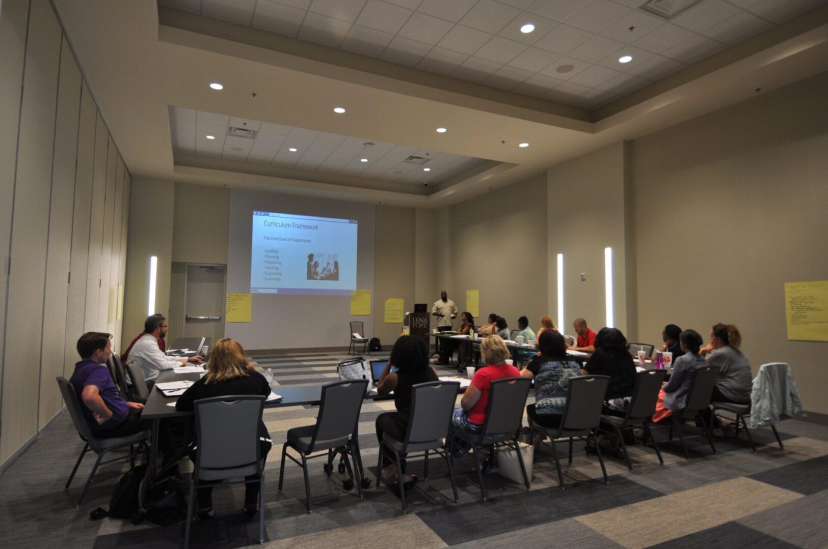 A group of people sitting around tables in front of a projector screen.