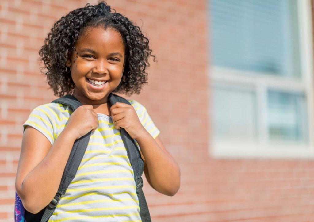 A young girl with curly hair is holding her backpack.